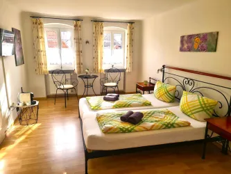 Ein gemütliches Schlafzimmer mit zwei Betten und bunten Kissen. Die Fenster lassen viel Licht herein und es gibt einen kleinen Tisch mit Stühlen.