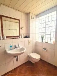 Ein modernes Badezimmer mit weißen Fliesen und einem großen Fenster. Es enthält ein Waschbecken, eine Toilette und einen Spiegel.