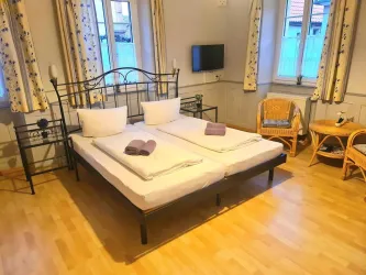Ein gemütliches Zimmer mit einem Doppelbett und Holzfußboden. Es gibt Fenster mit Vorhängen und einen Fernseher an der Wand.