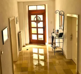 Ein heller Flur mit glänzenden Fliesen und einer offenen Eingangstür. Links steht ein Heizkörper und auf der rechten Seite ein kleines Möbelstück mit Zeitschriften.