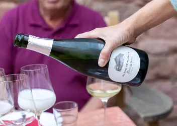 Eine Hand gießt einen Flascheninhalt in ein Glas. Im Hintergrund sind weitere Gläser und eine Person zu sehen.