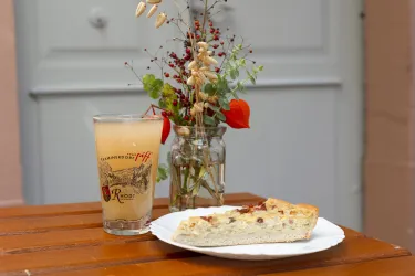 Ein Glas Getränk steht neben einem Stück Kuchen auf einem Teller. Im Hintergrund befindet sich eine kleine Vase mit frischen Blumen.