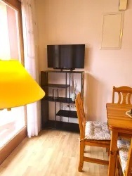 Ein gemütlicher Wohnraum mit einer Holztafel, einem Regal und einem Fernseher. Ein helles Licht steht auf dem Tisch und sorgt für eine einladende Atmosphäre.