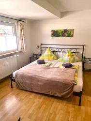 Ein gemütliches Schlafzimmer mit einem Doppelbett und bunten Kissen. Die Wände sind hell und es gibt ein Fenster mit Blick nach draußen.