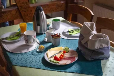 Ein gedeckter Tisch mit Frühstück, bestehend aus Wurst, Käse, frischem Gemüse und Getränken. Im Hintergrund befindet sich eine Kaffeekanne und frischer Orangensaft.