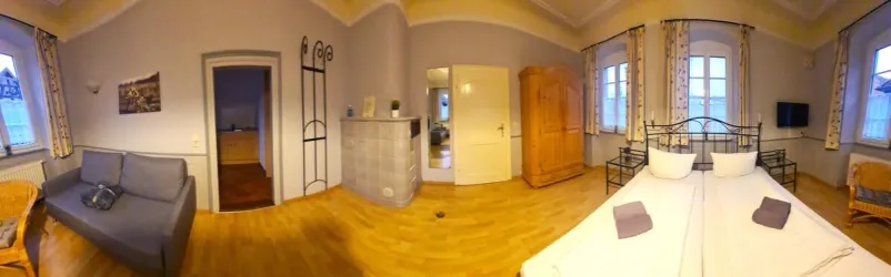 Ein helles Zimmer mit einem Doppelbett und einer Couch. Große Fenster sorgen für viel Tageslicht.