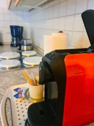 Eine moderne, rote Kaffeemaschine steht auf einem Tablett in einer Küche. Im Hintergrund sind Kochplatten und ein Wasserkocher sichtbar.