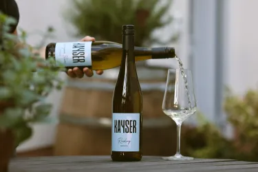 Eine Flasche Weißwein wird in ein Glas gegossen. Im Hintergrund sind Fässer und Pflanzen zu sehen, die eine gemütliche Atmosphäre schaffen.