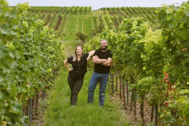 Ein Paar steht glücklich zwischen Weinreben in einem Weinberg. Im Hintergrund sind sanfte Hügel und weitere Rebstöcke zu sehen.