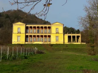 Ein gelbes historisches Gebäude mit Säulen und großen Fenstern. Es liegt in einer grünen Umgebung mit Bäumen im Hintergrund.