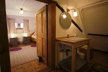 Ein gemütliches Zimmer mit Holzdetails und sanften Farben. Auf der linken Seite befindet sich ein Waschbecken und zur rechten Seite ein Blick auf ein Bett mit angeschlossenen Nachttischen.