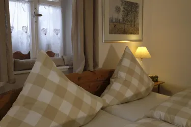 Ein gemütliches Schlafzimmer mit karierten Kopfkissen und einer warmen Tischlampe. Vor einem Fenster hängen helle Vorhänge mit Herzmotiven.