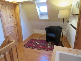 Ein gemütlicher Raum mit einem schwarzen Sessel und einem kleinen Tisch. Der Boden ist aus Holz und ein Teppich verleiht eine einladende Atmosphäre.