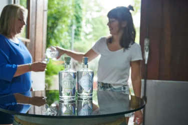Zwei Frauen stehen an einem Tisch mit Flaschen Gin. Eine Frau gießt ein Getränk in ein Glas.