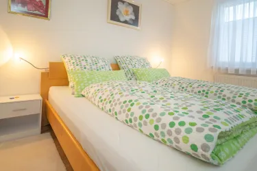 Ein gemütliches Schlafzimmer mit einem großen Bett und bunten Bettdecken. Es gibt eine Nachttischlampe und ein Fenster, das natürliches Licht hereinlässt.