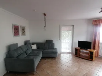 Ein gemütliches Wohnzimmer mit einem taubenblauen Sofa ausziehbar als Wohnlandschaft und einem Fernseher. Große Fenster lassen viel Licht herein.