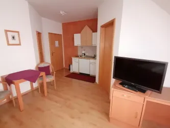 Ein helles Zimmer mit einer kleinen Küchenzeile und einem Fernseher. Es gibt einen Tisch mit Stühlen und eine warme, einladende Atmosphäre.
