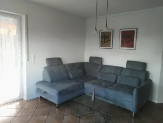 Ein modernes Wohnzimmer mit einer taubenblauen Couch ausziehbar und einem Glastisch. Die Wände sind hell und mit einigen Bildern dekoriert.