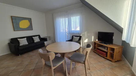 Ein gemütlicher Wohnbereich mit einem runden Tisch und Stühlen. Eine schwarze Ledercouch verwandelbar zu einem Bett. Ein Flachbild-TV ergänzen den Raum, der viel Tageslicht erhält. Integriert mit einer Küchenzeile.