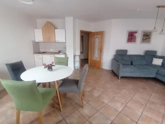 Ein modernes Wohnzimmer mit einer gemütlichen Couch und einem runden Esstisch. Der Raum hat helle Fliesen und eine offene Küchenzeile.