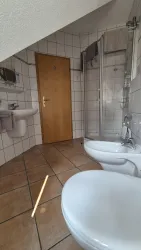 Ein helles Badezimmer mit einer Dusche, einem Waschbecken und einem WC. Der Boden ist mit Fliesen in warmen Tönen ausgelegt.
