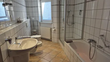 Ein modernes Badezimmer mit weißen Fliesen und einem großen Fenster. Es enthält eine Badewanne, eine Dusche, ein Bidet sowie Waschbecken und Toilette. Ausstattung mit Kosmetikspiegel und Föhn.