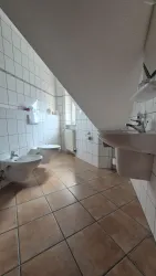 Ein modernes Badezimmer mit einem schrägen Dach. Es enthält ein Waschbecken, eine Toilette und einen Bidet, alles in hellem Fliesendesign.