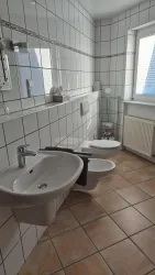 Ein modernes Badezimmer mit weißen Fliesen und einem Fenster, das natürliches Licht hereinlässt. Es gibt ein Waschbecken und eine Toilette.