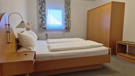 Ein einfaches Schlafzimmer mit einem Doppelbett und einem Nachttisch. An der Wand gibt es einen Schrank und ein Fenster mit Vorhängen.