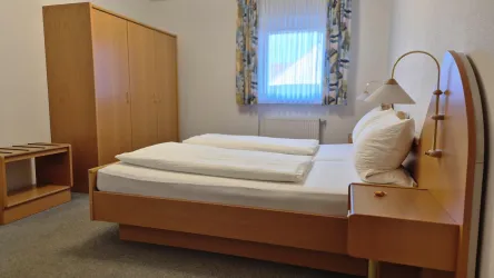 Ein einfaches Schlafzimmer mit einem Bett, das zwei Kissen und Decken hat. Es gibt einen Kleiderschrank und ein Fenster mit Vorhängen.