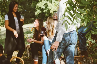 Eine Gruppe von Freunden sitzt im Freien und genießt gemeinsame Zeit. Sie lachen und halten Getränke in der Hand.