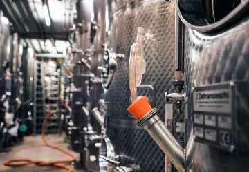 Eine moderne Weinproduktionsanlage mit glänzenden Edelstahltanks. Im Vordergrund ist ein Auslaufrohr mit einem orangefarbenen Deckel zu sehen.