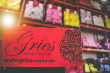 Ein rotes Schild mit dem Schriftzug „Gries“ und dem Slogan „Weine mit Identität“. Im Hintergrund sind Regale mit bunten Weinflaschen sichtbar.