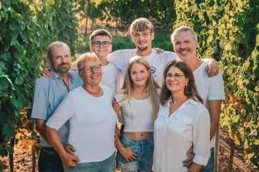 Eine Gruppe von sechs Personen steht zwischen Weinreben. Sie lächeln und tragen helle Kleidung.