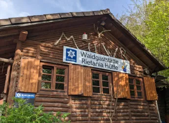 Ein gemütliches Holzhaus mit der Beschilderung „Waldgaststätte Rietania Hütte“. Die Fenster sind aus Holz und die Umgebung ist grün.