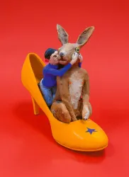 Ein gelber High Heel mit einer Figuren-Szene darin. Ein Mensch umarmt einen Hasen, während sie auf dem Schuh sitzen.