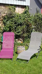 Zwei Liegen stehen auf einer grünen Wiese, eine lila und eine graue. Im Hintergrund sind Büsche und ein Holzfass zu sehen.