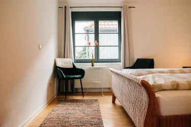 Ein helles Schlafzimmer mit einem Bett und einem Stuhl neben einem Fenster. Auf der Fensterbank steht eine Vase mit Tulpen.