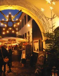 Eine festlich dekorierte Gasse mit Lichtern und Tannenbäumen in der Dämmerung. Besucher genießen die weihnachtliche Stimmung an einem Verkaufsstand.