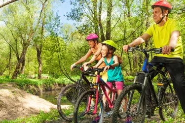 Eine Familie mit drei Radfahrern steht in der Natur. Sie tragen Helme und genießen die Zeit zusammen an einem kleinen Fluss.