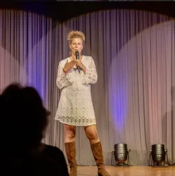 Eine Frau steht auf einer Bühne und spricht in ein Mikrofon. Sie trägt ein weißes Kleid und braune Stiefel, während der Hintergrund dezent beleuchtet ist.