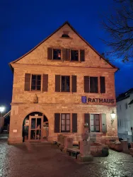 Ein historisches Rathaus mit Steinfassade und beleuchteten Fenstern. Der Abendhimmel ist dunkelblau, und der Platz vor dem Gebäude ist gepflastert.