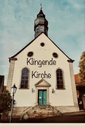 Eine historische Kirche mit einem markanten Turm und einem einladenden Eingang. Der Schriftzug "Klingende Kirche" ist prominent auf der Fassade zu sehen.