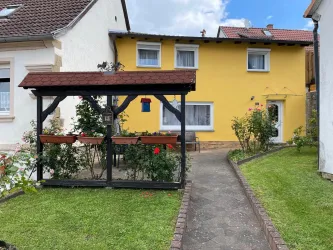 Ein gemütliches Haus mit gelber Fassade und einem kleinen Garten. Vor dem Haus steht ein pavillonähnlicher Bereich mit Rosen und Pflanzen.