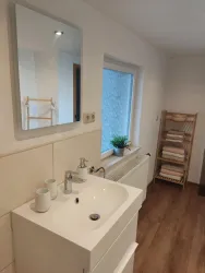 Ein modernes Badezimmer mit einem Waschbecken und einem großen Spiegel. An der Wand steht ein Regal mit Handtüchern und einer kleinen Pflanze.