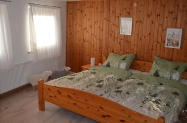 Ein gemütliches Schlafzimmer mit einem großen Holzbett und grünen Bettbezügen. Helle Vorhänge und eine Holzverkleidung verleihen dem Raum eine warme Atmosphäre.
