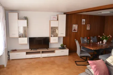 Ein modernes Wohnzimmer mit einem Fernseher auf einem niedrigen Regal. Auf der linken Seite ist ein Esstisch mit Stühlen, und die Wände sind teils hell und teils mit Holzverkleidung.