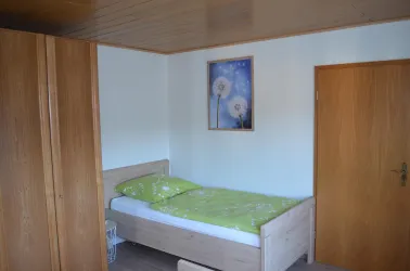 Ein einfaches Zimmer mit einem Einzelbett und einem grünen Bettlaken. An der Wand hängt ein Bild von Pusteblumen.