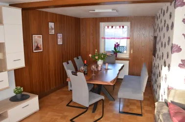 Ein gemütlicher Essbereich mit einem Holztisch und grauen Stühlen. An den Wänden hängen Bilder, und ein Fenster mit Blumen sorgt für Tageslicht.