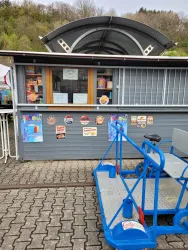 Ein Imbissstand mit einer großen Fensterfront und verschiedenen Speisekarten. Vor dem Stand steht ein blauer Anhänger.